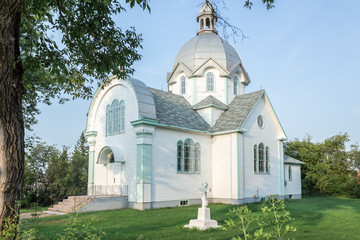 horizontal image of a huge big  white church with green trim and a concrete cross sitting on the yard with green grass and a big tree off to the side under a blue sky