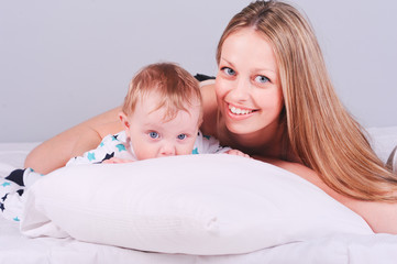 Beautiful mother lying with little child in bed. Smiling woman holding newborn child over gray. Motherhood