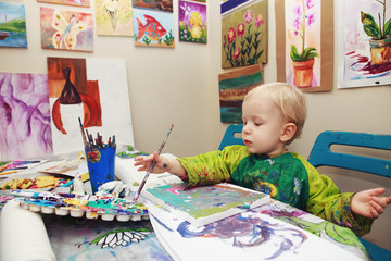 Fototapeta premium Cute adorable little baby boy girl toddler sitting in art studio indoors and drawing painting with brushes, pencils, pastel. Early child development concept