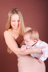Happy young woman playing with baby boy. Smiling stylish mother holding newborn child over brown. Motherhood
