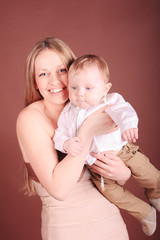 Happy young woman playing with baby boy. Smiling stylish mother holding newborn child over brown. Motherhood