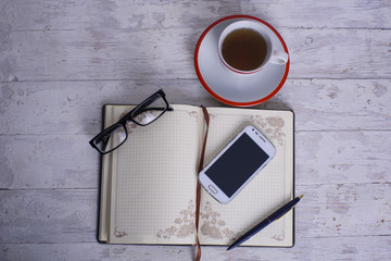 Naklejka premium cup, notepad, cellphone, glasses and pen on white vintage wooden background