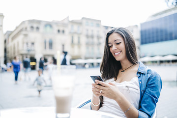 Young smiling woman with a drink on a terraced while sending tex