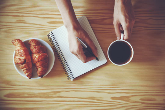 Hands with pen writing on notebook with morning coffee and crois