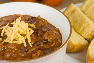Chili and Cornbread – A bowl of chili con carne topped with shredded cheddar cheese. Cornbread in the background.