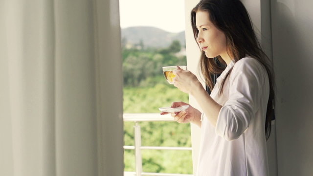 Young woman drinking tea standing by window at home, slow motion shot at 240fps
