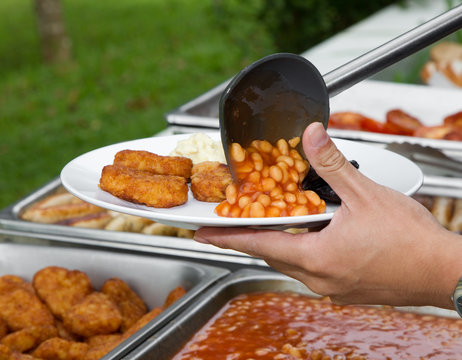 Hand serving a plate of baked beans, nuggets and mash potatoes with a plastic ladle