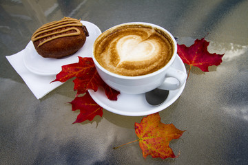 A hot coffee latte with foam in the shape of a heart and a chocolate peanut better dessert sit on a glass table with some red and orange leaves during autumn, Lake George, NY.