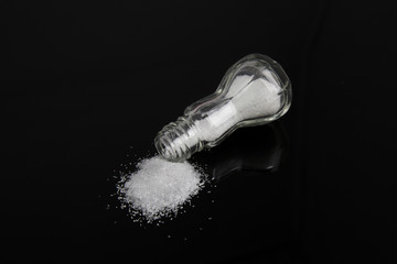 A salt shaker tipped over spilling salt on a reflective black surface with a black studio background.