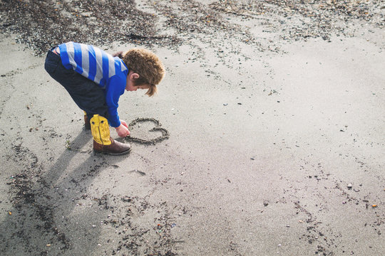 Boy in cowboy boots and raccoon hat drawing a heart in the sand