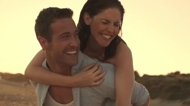 Young couple playing on beach in sunset, man carrying woman on his back.