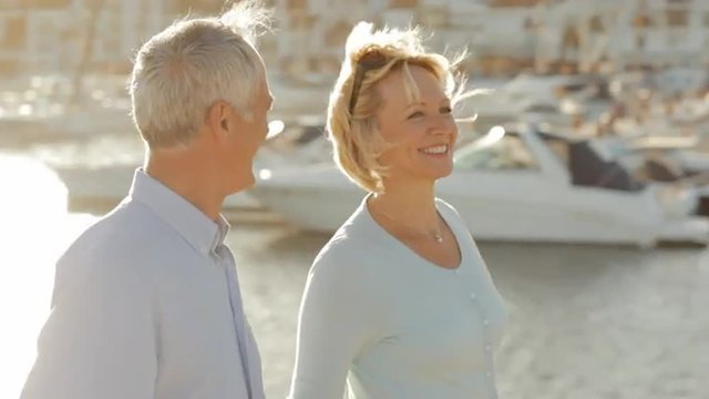 pan shot of senior couple walking by marina in sunset