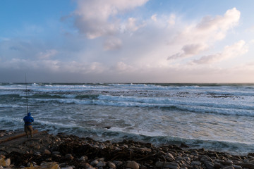 Fototapeta na wymiar Lone fisherman on a rocky, deserted beach under a stormy sky on the South African coast