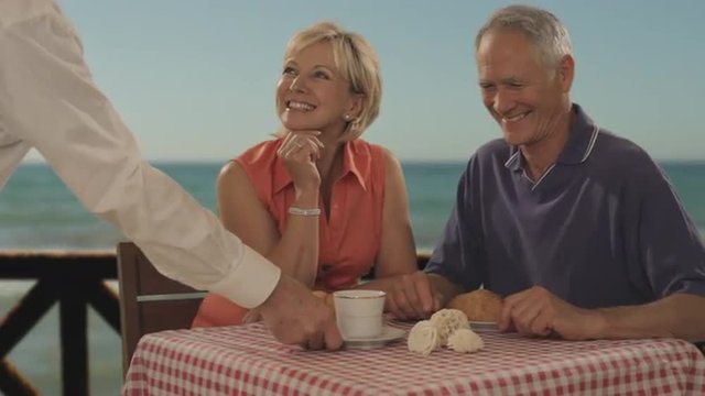 senior couple meeting at beach cafe by sea, waiter bringing coffee