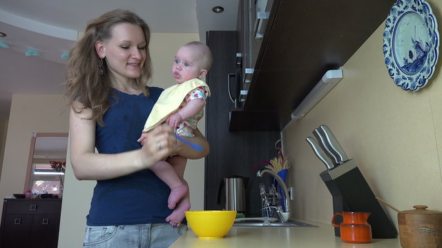 woman feed baby daughter with porridge holding on arm in kitchen