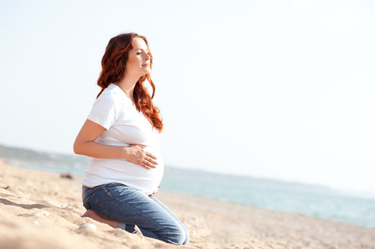 Resting pregnant girl sitting at beach outdoors. Holding belly over sea background. Motherhood. Maternity. 