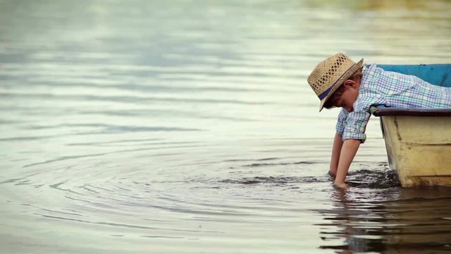 A little boy in the fancy hat dangling his hands in the water while on the boat
