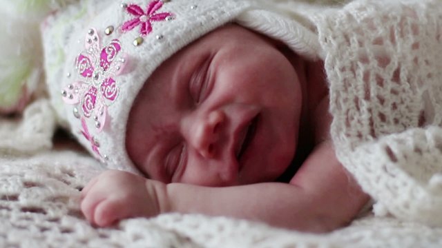 Little newborn baby in hat sleeps and grimaces on fluffy blanket
