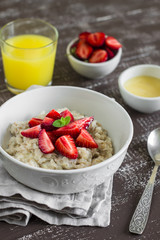 oatmeal with strawberries in a white bowl and orange juice on a dark wooden background