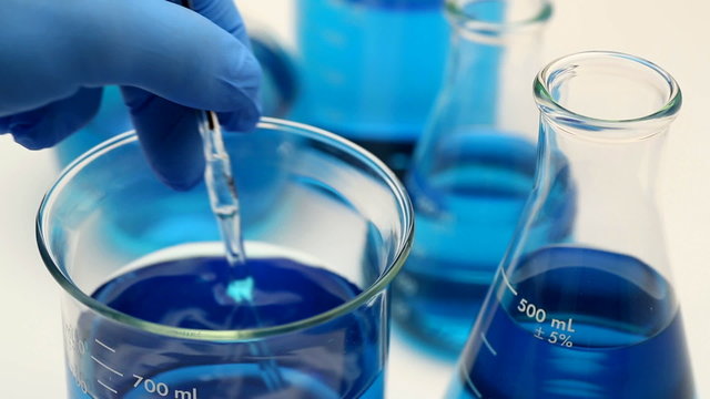 This is a close up photo of a scientist stirring a blue liquid in a beaker. He is wearing a blue rubber glove.