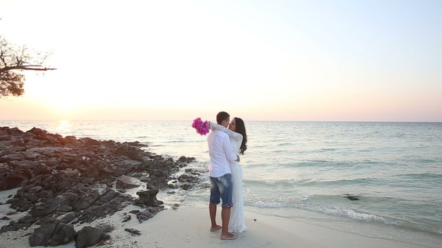 brunette bride and handsome groom barefoot hug at edge of water of beach at dawn