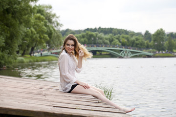Young beautiful girl in a white tunic sits on a wooden pier on t