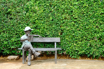 Boy reading on bench statue and green tree wall