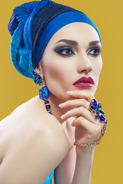 Beautiful middle eastern woman with hijab with blue scarf and blue jewelry and red lips on yellow background, studio shot. looking at camera. side looking.