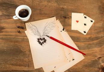 Tazzina di caffè con cuore di chicchi e ali disegnati,asso di cuori,cinque di picche e penna  su fogli di quaderno.