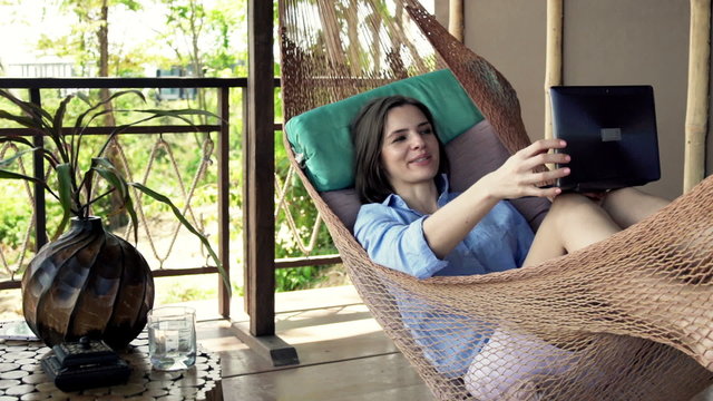 Young, happy woman chatting on laptop lying on hammock
