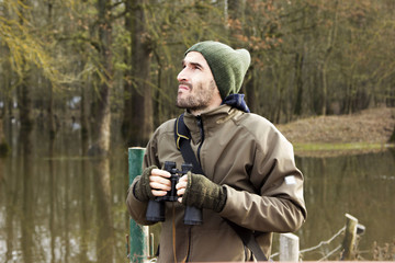 Man in jacket and binoculars standing on shore and observing river