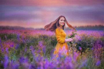 Beautiful young woman standing in the meadow of violet flowers at sunset.