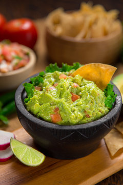 A delicious vertical shot Bowl of Guacamole next to fresh ingredients on a table with tortilla chips and salsa