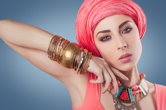 Beautiful fashion model with blue eyes with pink scarf (hijab) and gold jewelry looking at camera - studio shot, indian (middle eastern) style
on dark blue background.