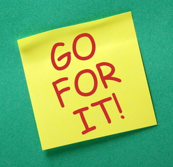 The phrase Go For It in red text on a yellow sticky note posted on a green notice board