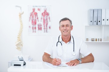 Smiling doctor writing on clipboard at his desk