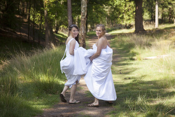 two brides walk on forest path with skirts in their arms