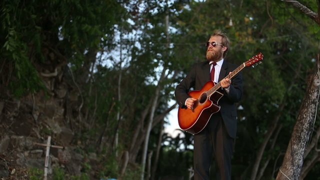 bearded guitarist in black suit makes up his mind to play against dark trees