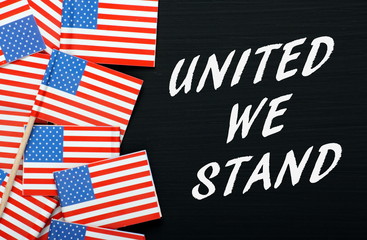 The phrase United We Stand in white text on a blackboard next to paper flags of the United States Of America