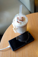 cell phone charging in the cafe with a plastic cup of iced chocolate frappe