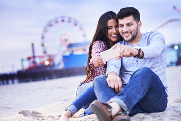 romantic couple sitting in sand taking selfie with santa monica pier in background