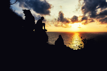 two human silhouettes sitting on a rock on the sunset ocean
