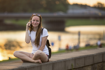 Young cute girl sitting on the promenade during sunset and talking on mobile phone.