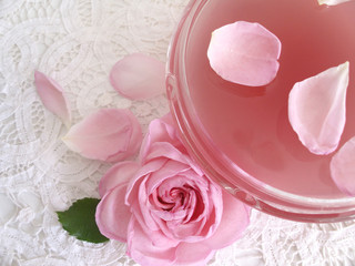 Kristallschale mit Rosenwasser gefüllt, daneben eine englische Rose und Rosenblätter, alles auf nostalgischem Deckchen.