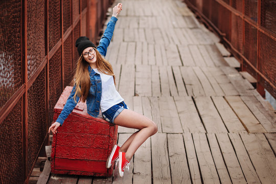Young sexy hipster woman sitting on red vintage box and laughing. Wearing jeans jacket, hipster black hat and glasses. Lifestyle city portrait.a