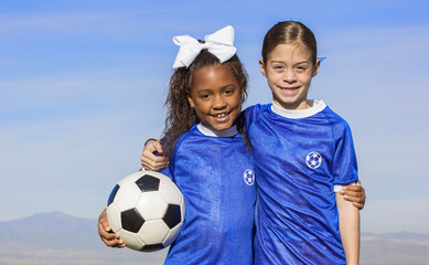 Cute, young african american and hispanic female soccer players holding a ball with a simple blue sky background - Powered by Adobe