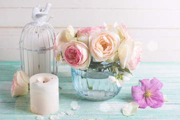 White roses, clematis and jasmine flowers  in vase and candles in ray of light  on turquoise wooden background against white wall. Place for text. Selective focus.