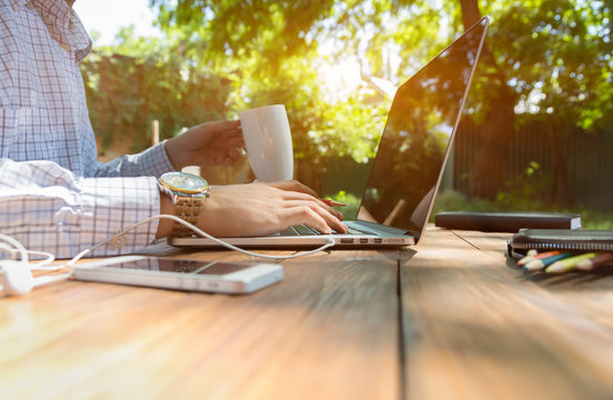 Escaped of office.
Business style dressed man sitting at natural country style wooden desk with electronic gadgets around working on laptop drinking coffee sunlight and green terrace on background