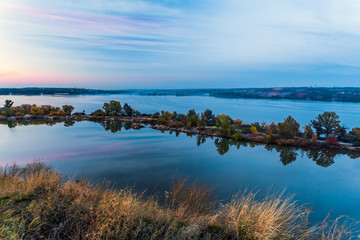 Tenderly colored sunset landscape at East European waterfront. Majestic view large river with unusual shape island in the middle small town buildings on remote waterfront soft color rose red dusk sky