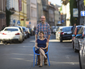 Couple, happy young family. Conceptual photo, a young woman sits on a chair in the middle of the street, a man stands behind her. European city blur in the background.